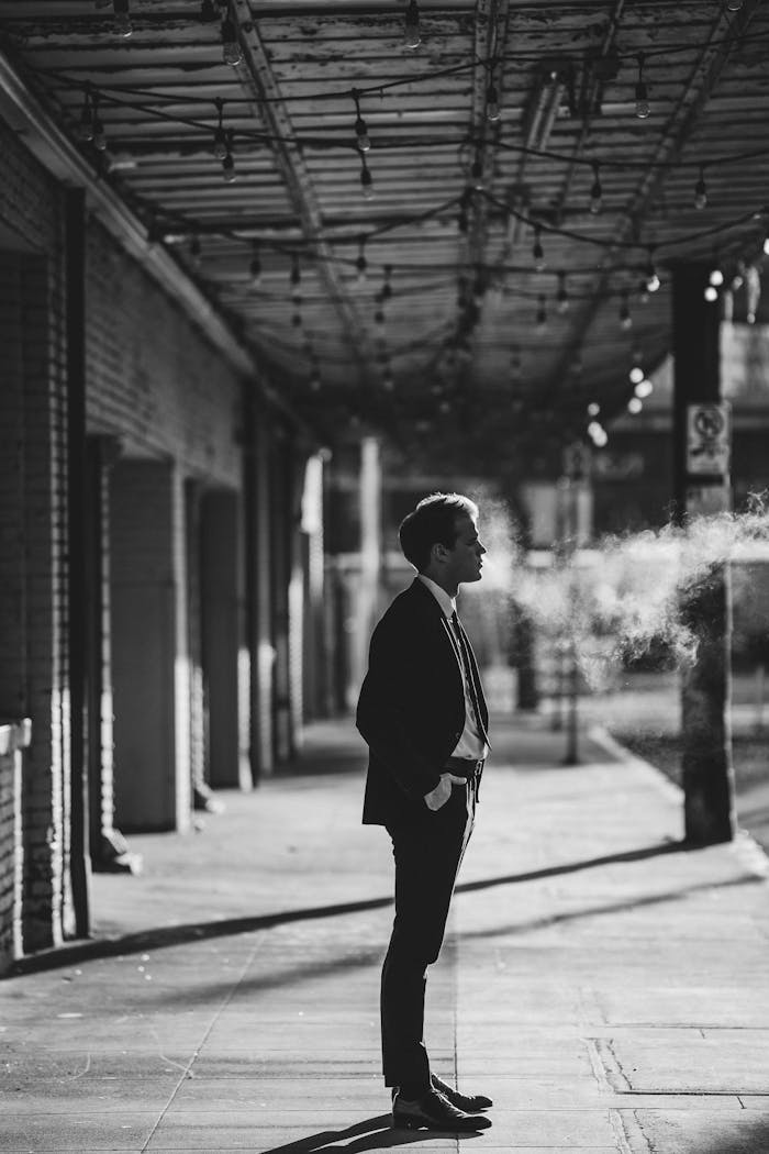 Black and white side view of male entrepreneur in formal suit with hand in pocket looking forward between haze and building on pathway